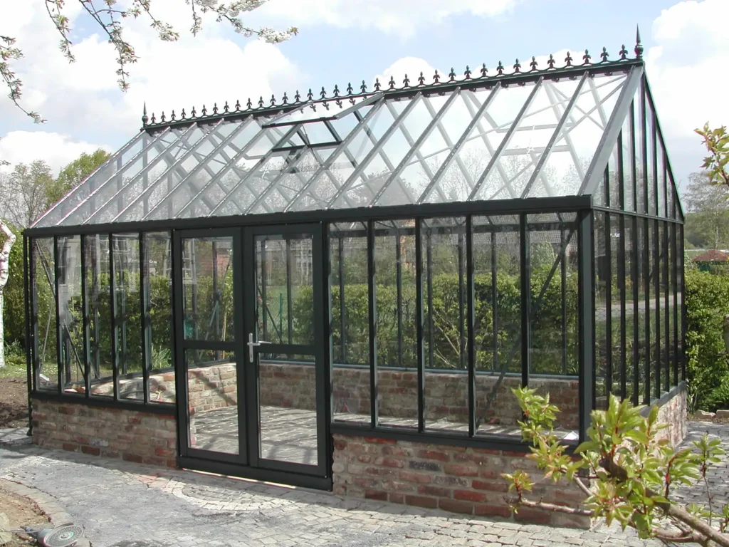 Glasgewächshaus mit Ziegelbasis von HOGATEK von Hof und Garten Wittstock. Gartenszene im Hintergrund.